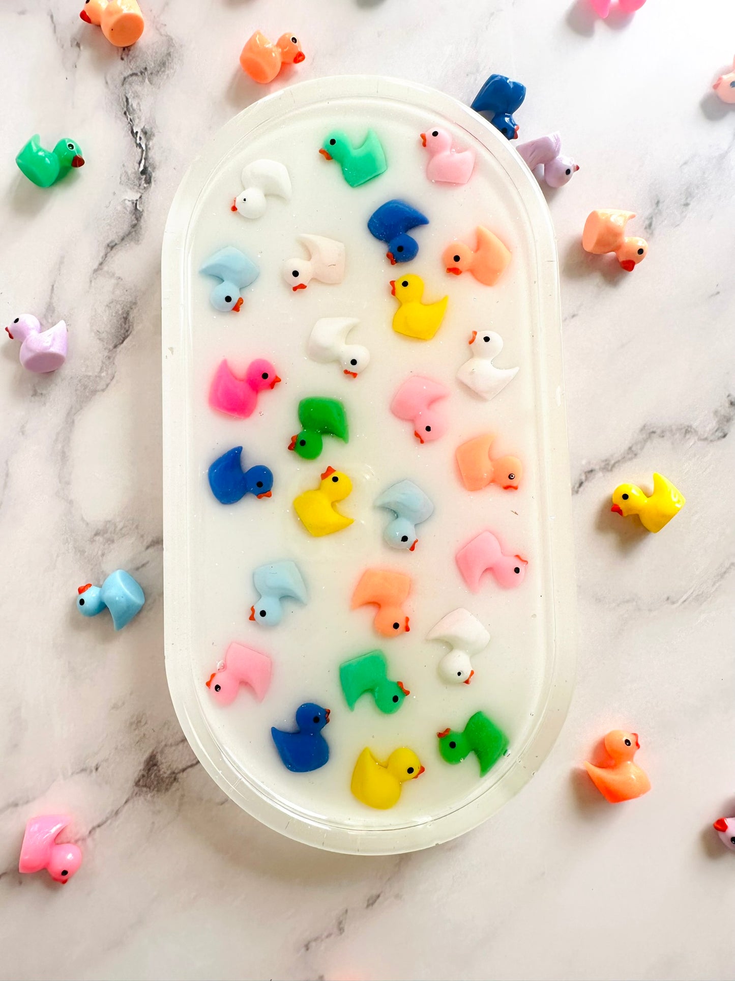 Oval-shaped resin tray with a glossy white base, featuring small colorful rubber duck figures embedded throughout. The ducks are arranged in neat rows and come in bright shades of pink, yellow, blue, green, orange, and white, giving the piece a playful and cheerful look against the marble background.