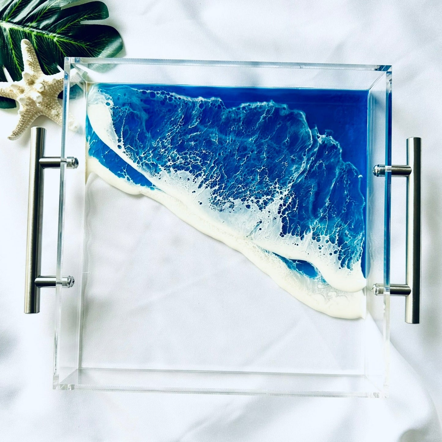 A clear acrylic serving tray with a blue ocean wave design, placed on a white surface with a shell and a green leaf for decoration.