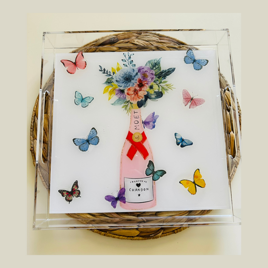 A decorative resin serving tray with a floral watercolor design and butterfly motifs, resembling a bottle of champagne, with a black and gold bow on top, placed on a wicker surface.