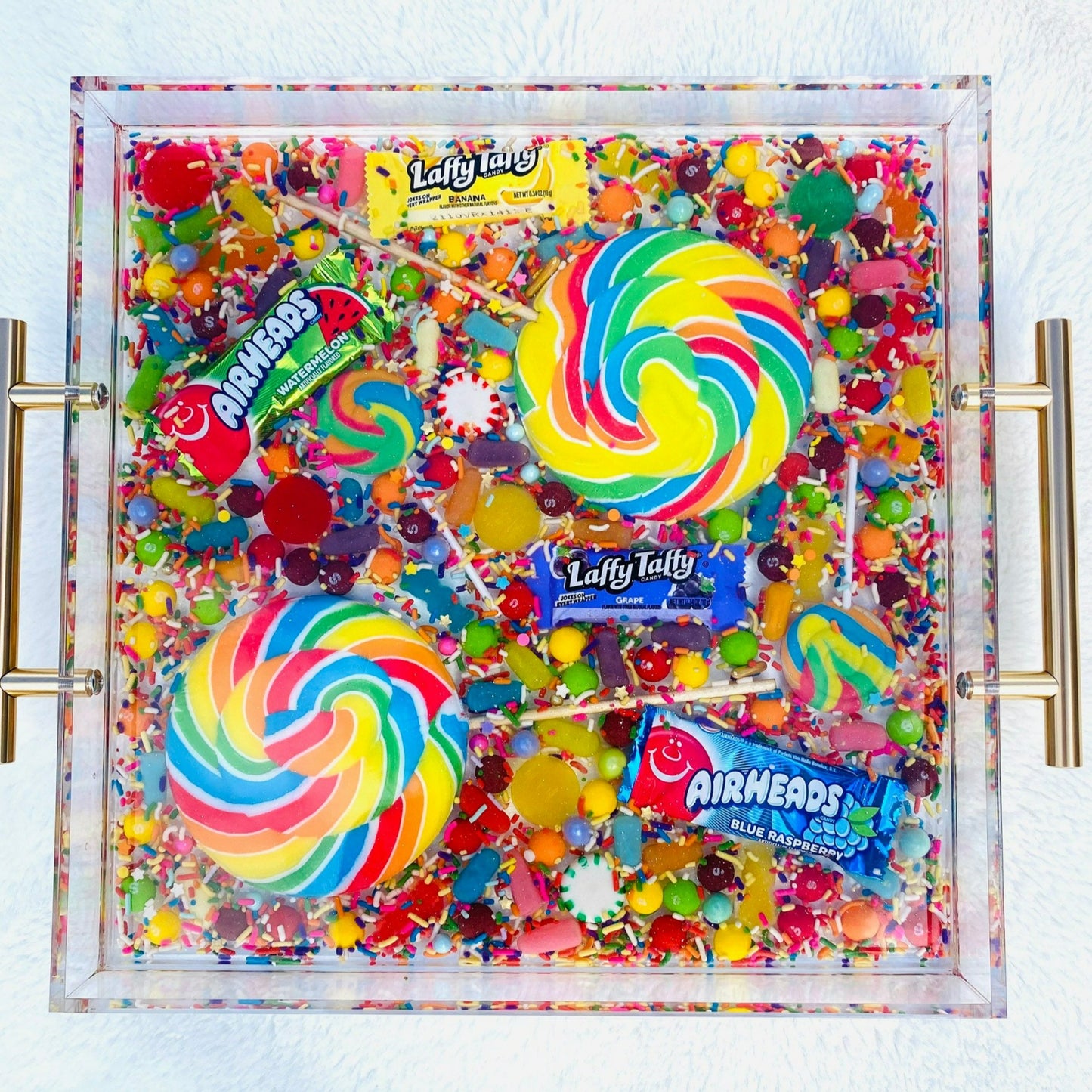 An acrylic serving tray filled with an assortment of colorful candy, including large and small pinwheel lollipops, and wrapped candies. The tray has a glossy finish with gold-tone handles.
