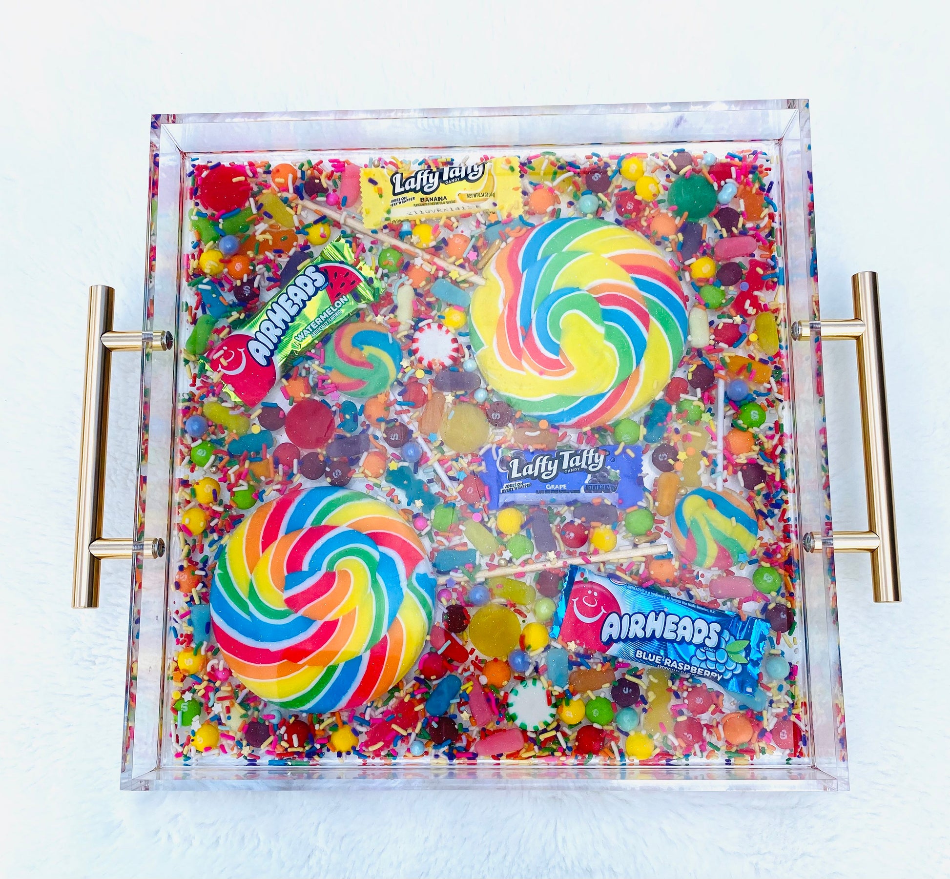 An acrylic serving tray filled with an assortment of colorful candy, including large and small pinwheel lollipops, and wrapped candies. The tray has a glossy finish with gold-tone handles.