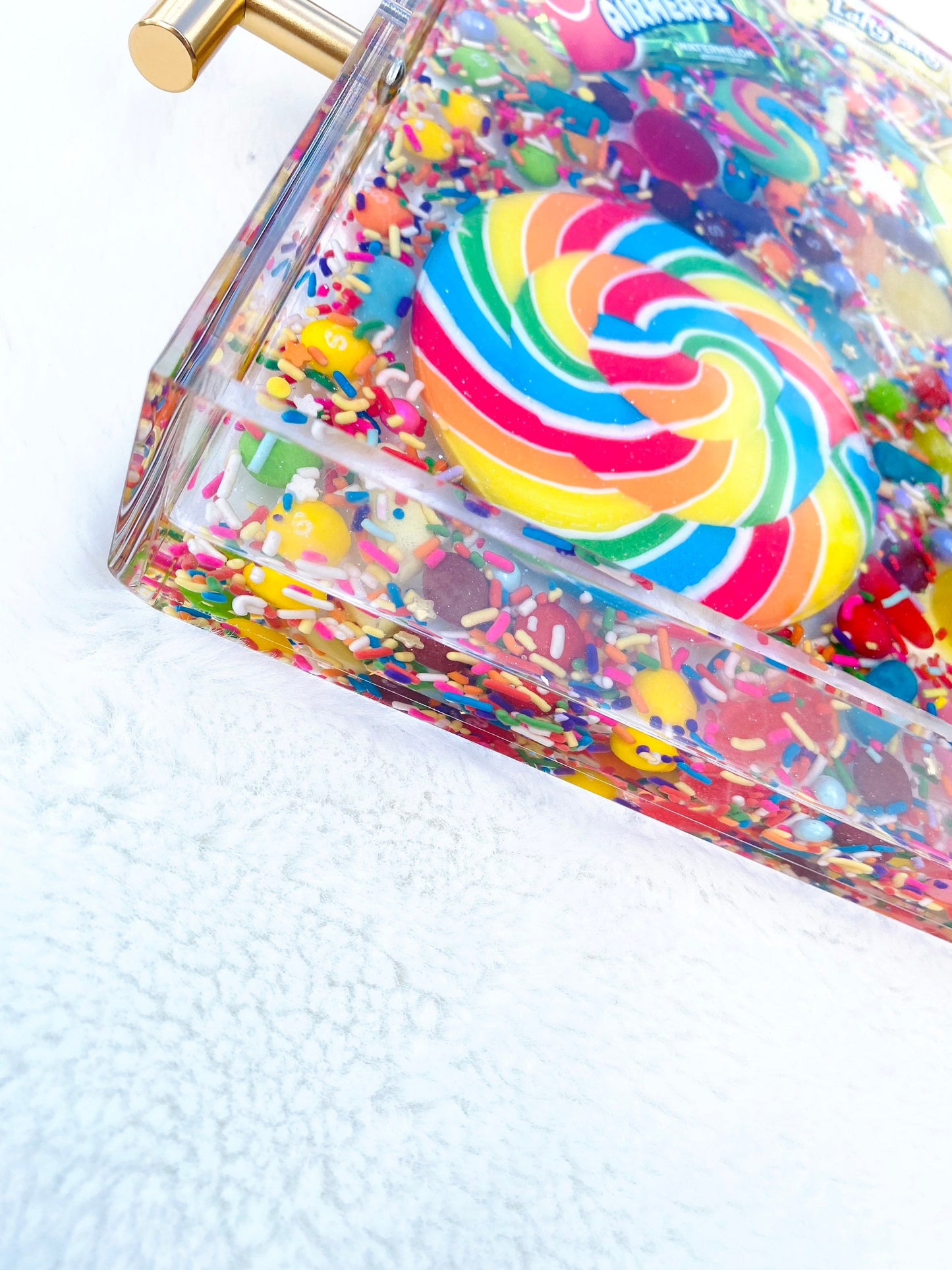 An acrylic serving tray filled with an assortment of colorful candy, including large and small pinwheel lollipops, and wrapped candies. The tray has a glossy finish with gold-tone handles.