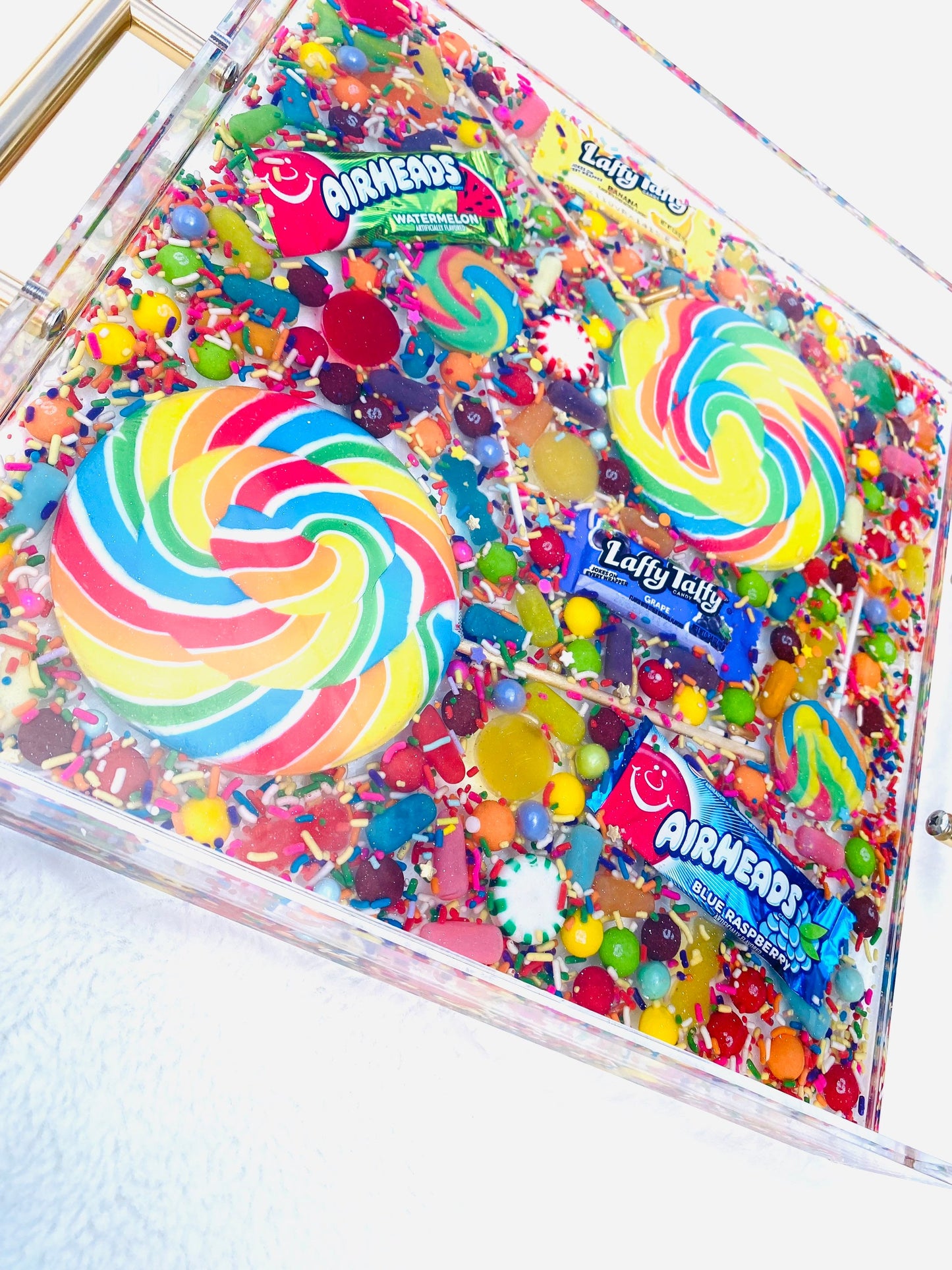 An acrylic serving tray filled with an assortment of colorful candy, including large and small pinwheel lollipops, and wrapped candies. The tray has a glossy finish with gold-tone handles.