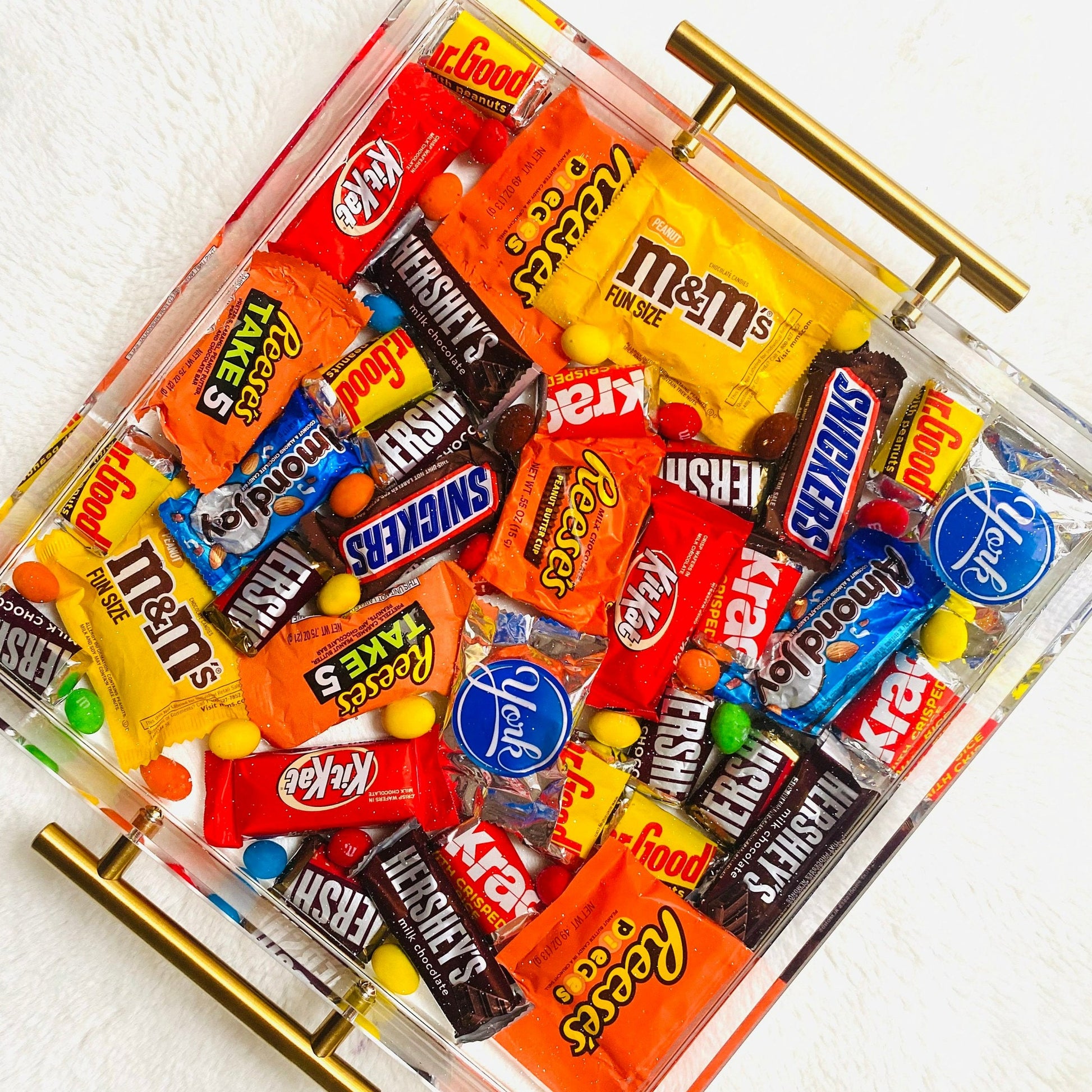 An acrylic serving tray filled with an assortment of chocolate candies, including brands such as M&M's and reeses, with a glossy finish and gold-colored handles.