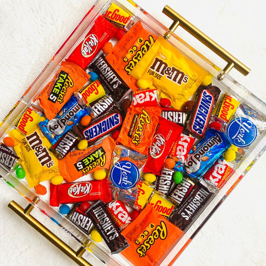 An acrylic serving tray filled with an assortment of chocolate candies, including brands such as M&M's and reeses, with a glossy finish and gold-colored handles.