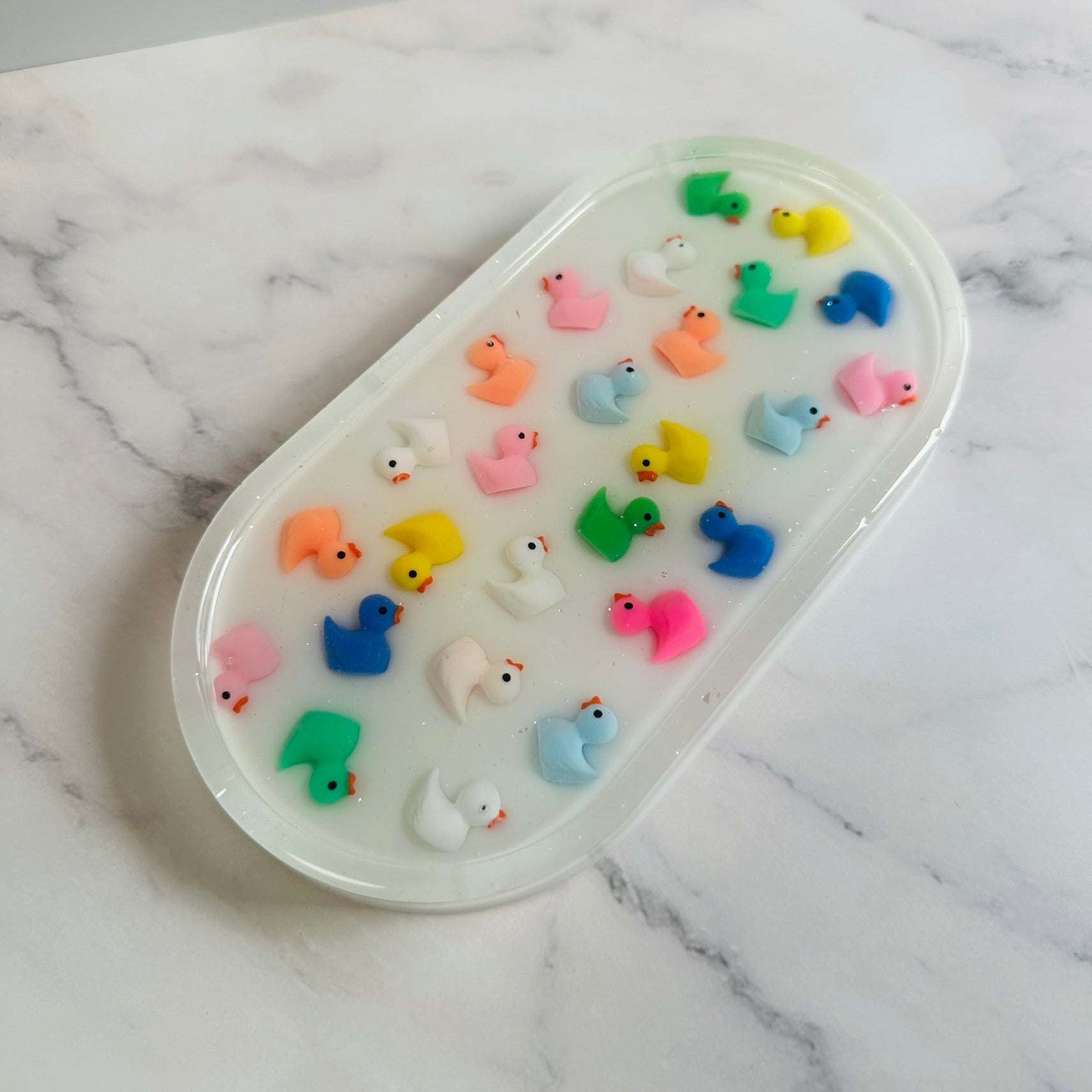 Oval-shaped resin tray with a glossy white base, featuring small colorful rubber duck figures embedded throughout. The ducks are arranged in neat rows and come in bright shades of pink, yellow, blue, green, orange, and white, giving the piece a playful and cheerful look against the marble background.