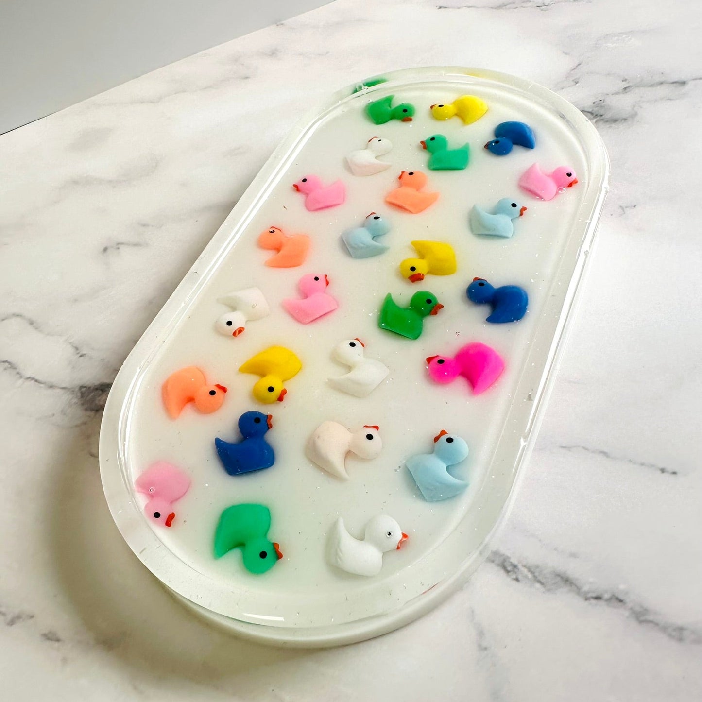 Oval-shaped resin tray with a glossy white base, featuring small colorful rubber duck figures embedded throughout. The ducks are arranged in neat rows and come in bright shades of pink, yellow, blue, green, orange, and white, giving the piece a playful and cheerful look against the marble background.