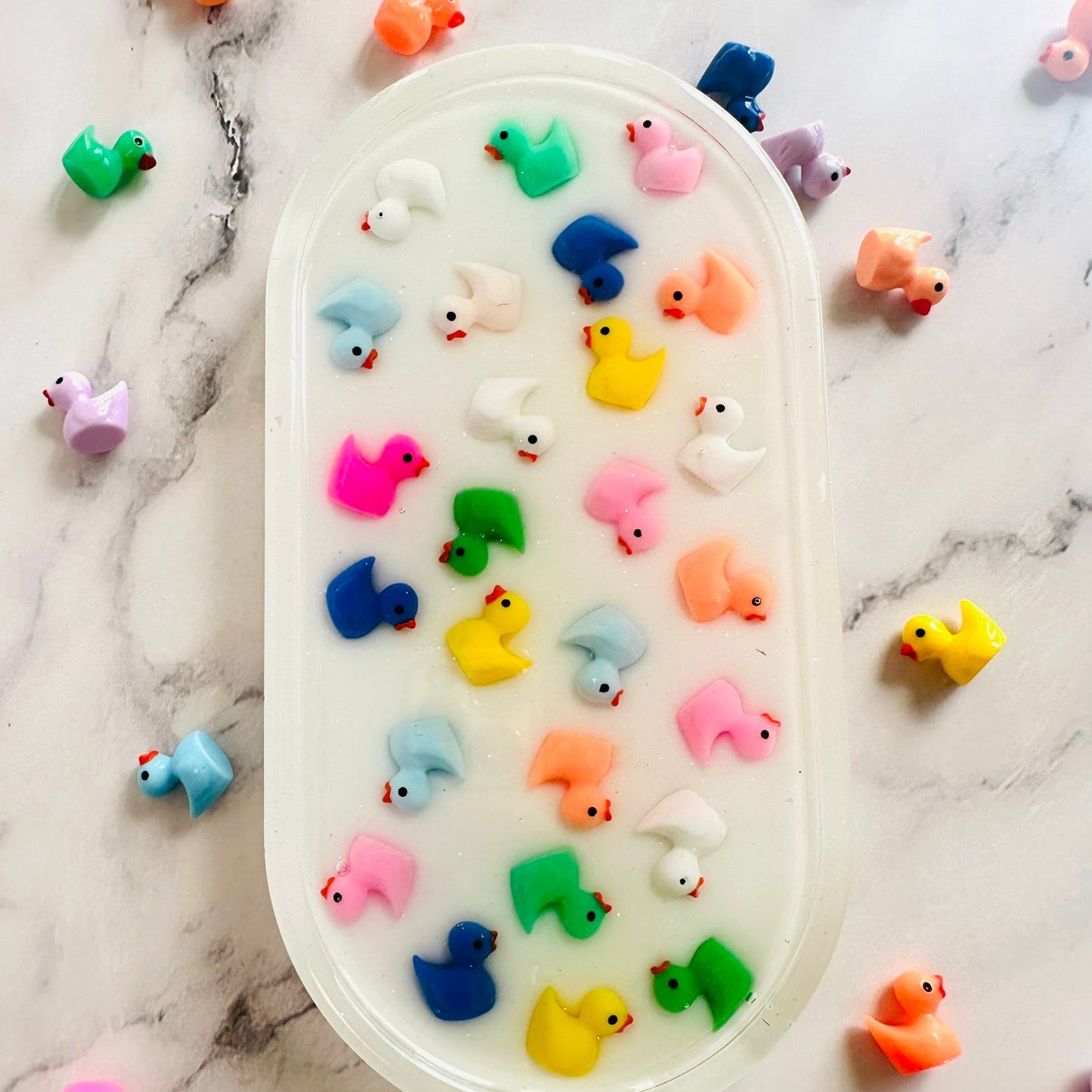 Oval-shaped resin tray with a glossy white base, featuring small colorful rubber duck figures embedded throughout. The ducks are arranged in neat rows and come in bright shades of pink, yellow, blue, green, orange, and white, giving the piece a playful and cheerful look against the marble background.